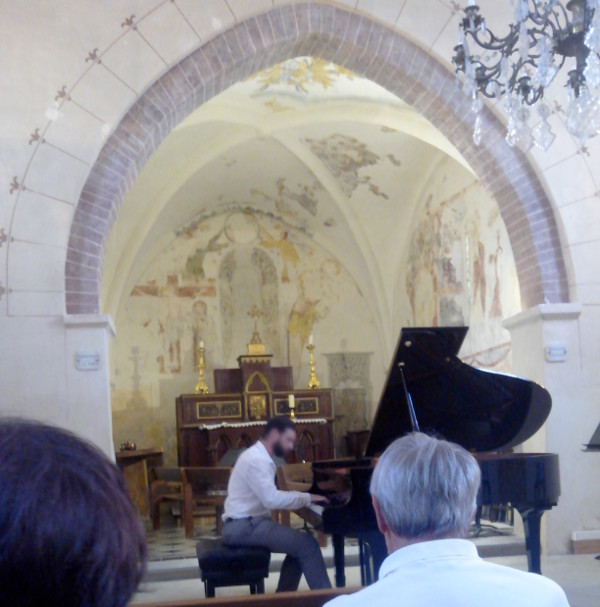 Vue de l'int&eacute;rieur de l'&eacute;glise, on aper&ccedil;oit un lustre en haut &agrave; droite, au centre il y a un pianiste (au visage flout&eacute;), et sur le devant, des spectateurices de dos.